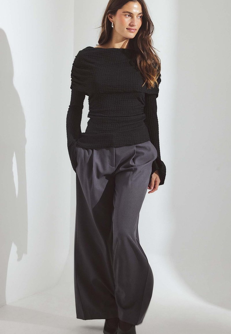 Čierne textúrované tričko s dlhým rukávom a lodičkovým výstrihom, skombinované s vysokými šedými nohavicami so širokými nohami. Model stojí na svetlom pozadí.