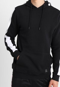 Svart hoodie i mjukt tyg med en framficka, justerbara snoddar och vita detaljer på ärmarna. Ribbad mudd och nederkant.