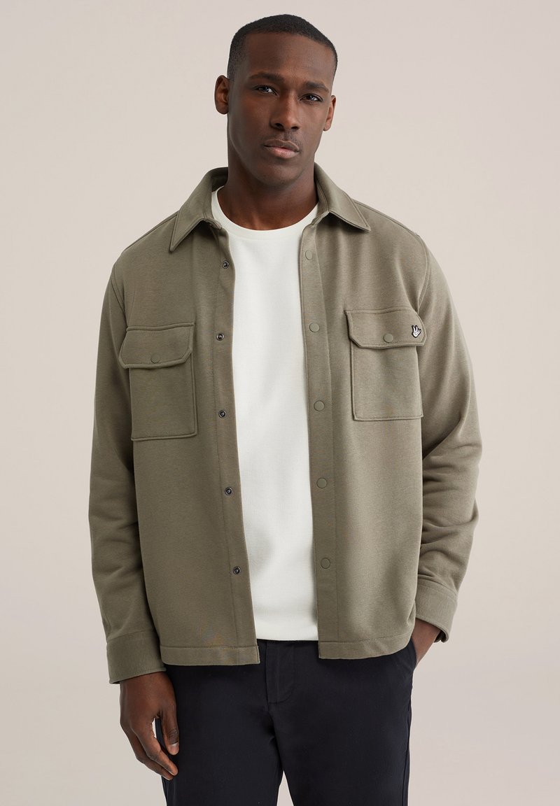 Olivo zelena jakna s srajčoim krojem, narejena iz mehke tkanine, z ovratnikom, dvema žepoma na prsih in pritiskači. Nošena čez belo majico.