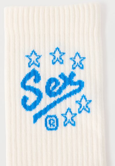 Λευκό ύφασμα με ραβδώσεις και μπλε πλεκτό κείμενο "See" υπογραμμισμένο, περιτριγυρισμένο από έξι μπλε αστέρια και ένα μικρό σύμβολο καταχωρημένου εμπορικού σήματος.