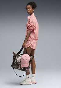 Ροζ νάιλον μπουφάν και σορτς, κρατώντας μια σκουρόχρωμη τσάντα με σχέδια και ροζ καπέλο. Λευκά αθλητικά παπούτσια με ροζ λεπτομέρειες και λευκές κάλτσες.