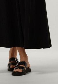 Sorte lædersandaler med to brede stropper, en spænde detalje, og en kraftig sål. Positioneret under en flydende sort nederdel.