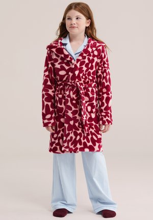 Fille portant un peignoir à motif floral rouge et rose par-dessus un pyjama rayé bleu et blanc, debout sur un fond clair uni.