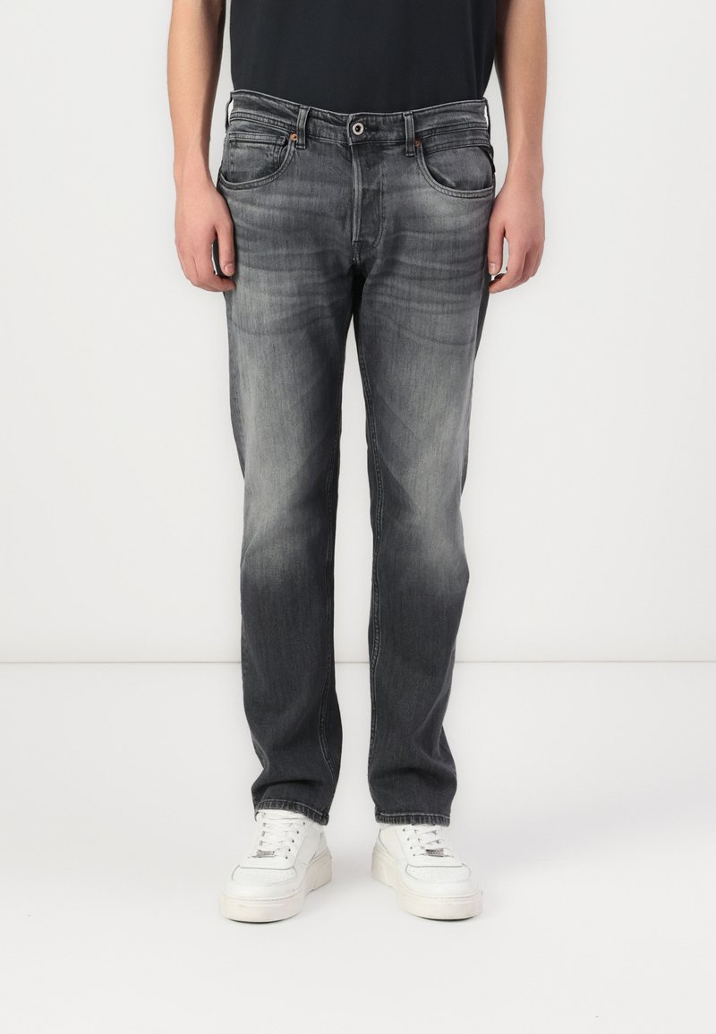 Blugi din denim gri cu un finisaj decolorat, două buzunare frontale și închidere cu buton, asortați cu teniși albi și o cămașă neagră.