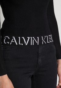 Svart ribbad tröja med ett brett elastiskt midjeband med vit "CALVIN KLEIN"-text, ihop med svarta jeans, som visar en figurnära form.