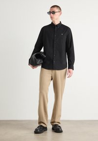 Black button-up shirt with a small logo, beige trousers, and black shoes. Holding a black bag. Model wears dark sunglasses.