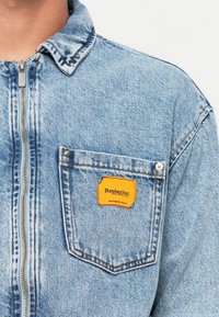 Light blue denim jacket with a front zipper and chest pocket featuring a yellow "Replay Inc." label.