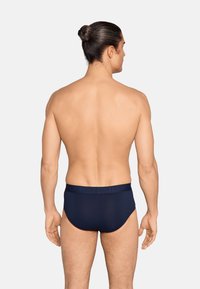 Homme aux cheveux attachés, torse nu, portant un slip bleu marine, debout de dos sur un fond uni.