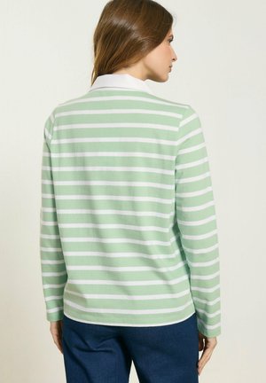 Frau, die ein langärmeliges, hellgrün-weiß gestreiftes Shirt mit weißem Kragen trägt, Ansicht von hinten.