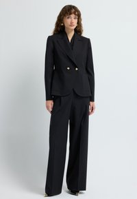 Zwarte op maat gemaakte pak met een dubbelzijdige blazer met gouden knopen en wijde broekspijpen. Gladde stof en gestructureerd ontwerp.