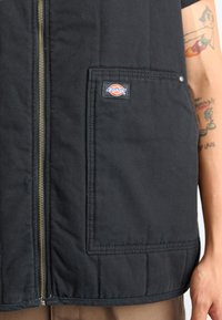 Črna brezrokavnika z zadrgo spredaj, izdelana iz trpežne tkanine z zaklopnim žepom. Ima zlato zadrgo in večbarvno oznako blagovne znamke, prišito na oblačilo.