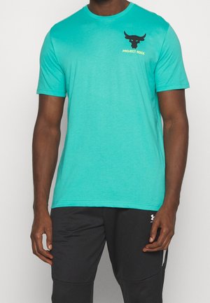 Mężczyzna w turkusowej koszulce Project Rock i czarnych spodniach Under Armour, stojący na jednolitym białym tle.