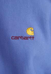 Fialová tkanina s ribovanou texturou, na které je vyšitý zlatý logo a červený text "carhartt." Zaměřte se na charakteristické víření loga.