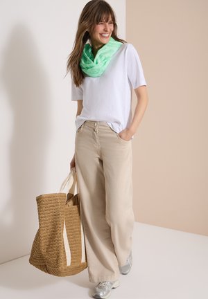 Nainen, jolla on valkoinen t-paita, beiget leveälahkeiset housut, vihreä huivi, hopeiset lenkkarit ja suuri kudottu laukku, hymyillen ja kävellen.