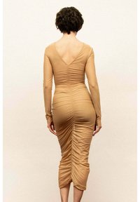 Sukienka przylegająca do ciała, z długim rękawem, w jasnobrązowym kolorze. Posiada wycięcie w kształcie litery V na plecach, marszczenia po bokach oraz długość sięgającą do połowy łydki.