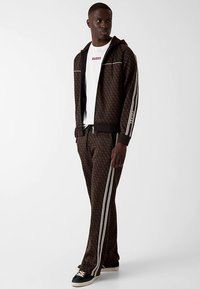 Brauner Trainingsanzug mit Muster und weißen Streifen. Besteht aus einer Kapuzenjacke mit Reißverschluss und passenden Hosen. Verziert mit einem Logo und weißen Details.