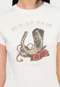 Lys farget t-skjorte med grafisk trykk av en hestesko, cowboystøvel, tau og røde roser, med teksten "TOMMY JEANS" tydelig vist.