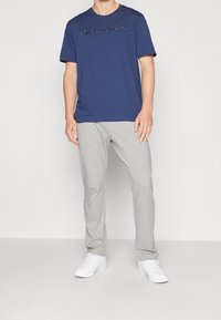 Námořnické tričko s krátkým rukávem s vyšitým logem "Champion", spárované se světle šedými tepláky a bílými teniskami. Hladké pozadí.