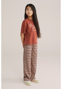 Brūns īsām piedurknēm t-krekls ar "Rosa" grafisku tekstu, kombinēts ar vaļīgām sarkanām paisley raksta biksēm un bēšām kurpēm, stāvot uz neitrāla fona.