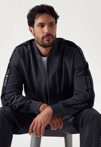 Black bomber jacket with zip front, ribbed cuffs, and embossed "GUESS" logo on sleeves. Worn over a white shirt, styled casually.