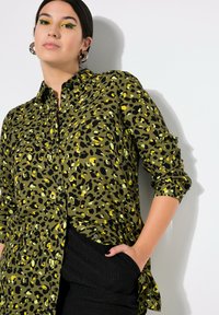 Blusa de estampado de leopardo verde con acentos en negro y amarillo, de manga larga, con cierre de botones y un corte relajado. Combinada con pantalones negros texturizados.