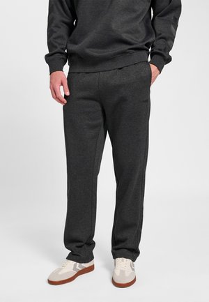 Man draagt donkergrijze sweatpants en bijpassend sweatshirt, staat met handen in de zakken, witte en bruine sneakers zichtbaar.
