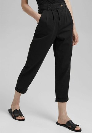 Femme portant un pantalon noir taille haute fuselé avec des ourlets retroussés et des sandales noires faciles à enfiler, debout avec une main dans la poche.