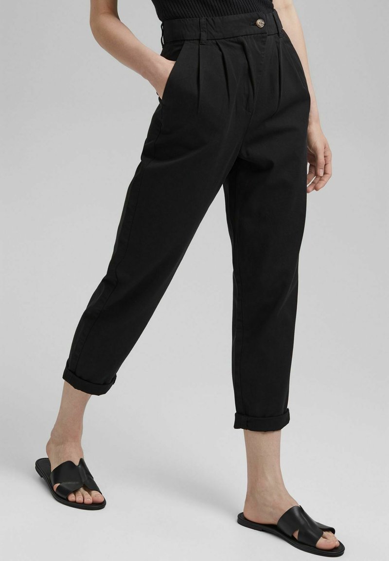 Femme portant un pantalon noir taille haute fuselé avec des ourlets retroussés et des sandales noires faciles à enfiler, debout avec une main dans la poche.