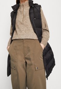 Svart pufferväst med hög krage och dragkedja, lager-på-lager över en beige stickad tröja, kombinerad med khaki vida byxor med sidofickor.