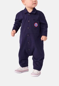 Marineblå bomulls jumpsuit med lange ermer, knappefront og brystlomme med et sirkulært brodert logo. Hvite sneakers.