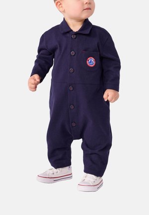 Navy-blaues Baumwoll-Overall mit langen Ärmeln, Knopfleiste vorne und Brusttasche mit einem runden gestickten Logo. Weiße Sneakers.