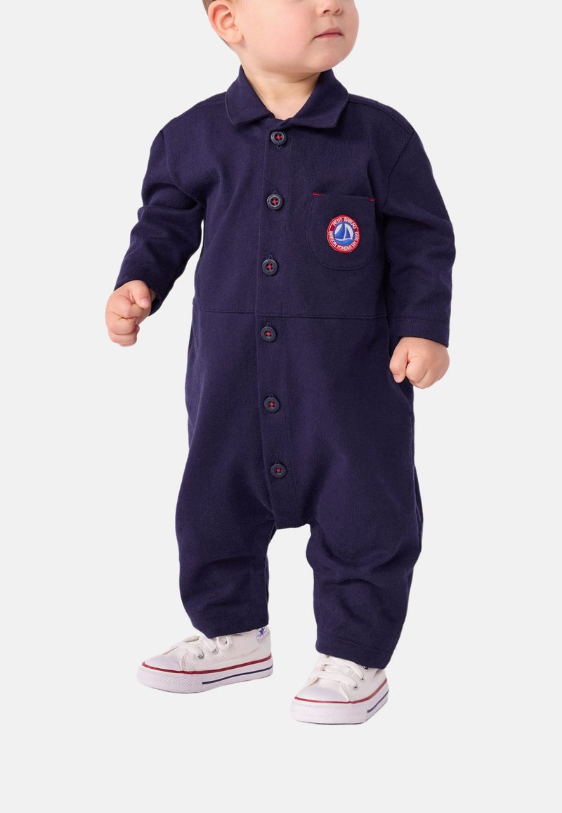 Marineblå bomulls jumpsuit med lange ermer, knappefront og brystlomme med et sirkulært brodert logo. Hvite sneakers.