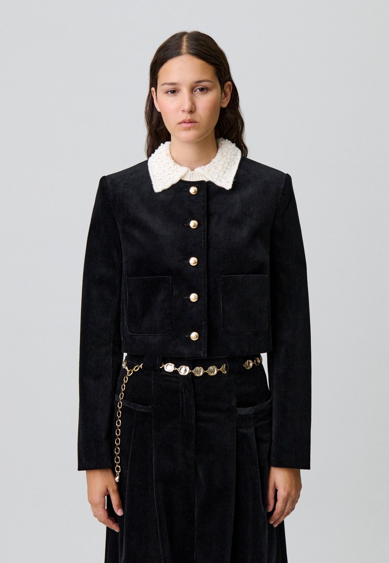 Giacca cropped in corduroy nera con colletto in maglia bianca, dotata di bottoni dorati e due tasche anteriori. Abbinata a una gonna coordinata e a una cintura.