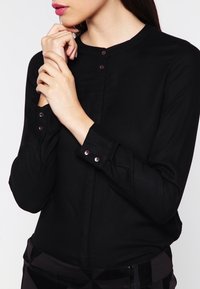 Black button-up blouse with a round neckline, long sleeves, and two front buttons, featuring a smooth fabric texture and fitted design.