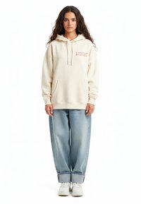 Felpa a cappuccio color crema con tasca frontale e testo ricamato. Abbinata a jeans baggy in denim chiaro e sneakers bianche.