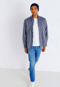 Karierte Hemdjacke in Marineblau und Weiß über einem weißen T-Shirt, kombiniert mit blauen Jeans und weißen Sneakern; trägt eine Marineblaue Mütze.