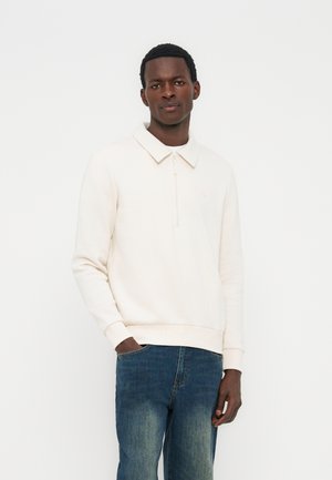 Mladý muž s tmavou pokožkou, oblečený v bielom pulóvri s dlhým rukávom, golierom a zipsom, v kombinácii s modrými džínsami, stojaci na bielom pozadí.