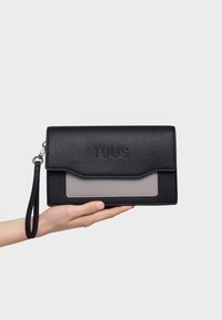 Black textured leather clutch with a gray panel, embossed "TOUS" logo, wrist strap, and flap closure, held in a hand against a white background.