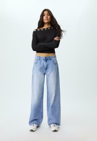 Donna con lunghi capelli scuri, in piedi con le braccia incrociate, indossa un maglione nero corto con occhielli metallici e jeans a gamba larga di un blu chiaro.