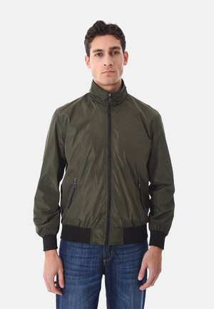 Uomo che indossa una giacca leggera verde oliva con cerniera, polsini e fascia in vita neri, in piedi davanti a uno sfondo chiaro e uniforme.