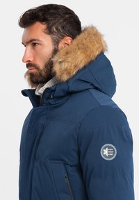 Marinblå puffjacka med huva klädd i fuskpäls. Har dragkedjefickor och ett cirkulärt emblem på ärmen. Tjockt, isolerat tyg.