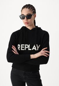 Hoodie noir en tricot avec "REPLAY" en lettres argentées, associé à des lunettes de soleil noires angulaires, modèle avec les bras croisés.