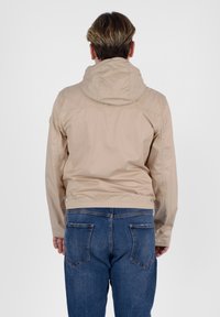 Giacca leggera beige con cappuccio, che mostra una texture liscia e polsini elasticizzati, abbinata a jeans in denim blu con tasche posteriori.