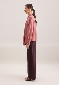 Blusa rosa chiaro con maniche a sbuffo, abbinata a una lunga gonna bordeaux scuro. L'outfit presenta un design semplice e una texture liscia.