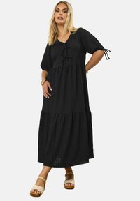 Vestido largo negro con mangas cortas abullonadas, que presenta un escote cuadrado, detalle de cordón y falda con volantes. Llevado con sandalias de plataforma color marrón.