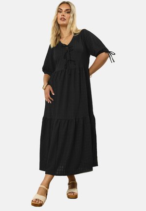 Schwarzes Maxikleid mit kurzen Puffärmeln, das einen quadratischen Ausschnitt, einen Kordelzug und einen gestuften Rock aufweist. Getragen mit beigen Plateau-Sandalen.