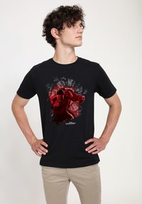 Camiseta de algodón negra con un diseño gráfico rojo y negro de un personaje musculoso con efectos explosivos detrás de él. Mangas cortas.