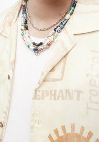 Collane di perline sovrapposte a una camicia bianca e una camicia leggero a maniche corte con motivo. Le collane presentano perline multicolori e accenti in argento.