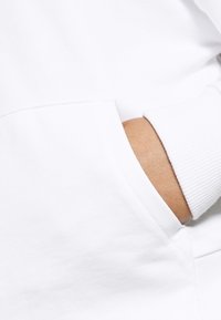 Hoodie bianco con tasca frontale; tessuto morbido, polsini a coste e cuciture visibili. La tasca mostra una mano all'interno, evidenziando una texture della pelle liscia.