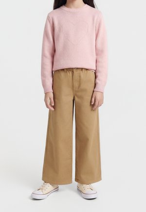 Barn iført en lyserød sweater med hjerte mønster, beige vidde bukser og cremefarvede sneakers, der står mod en hvid baggrund.